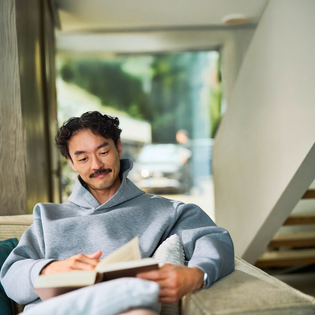 Un cliente rilassato che usufruisce del servizio assistenza mobile a casa propria Un cliente seduto in casa intento a leggere un libro. Da una finestra alle sue spalle si vede un'auto durante la manutenzione effettuata da un tecnico.